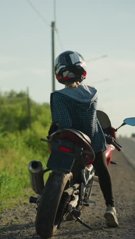 una vista trasera de una motociclista femenina, relajada en su bicicleta, mirando contemplativamente a la izquierda y a la derecha en una carretera serena con vegetación exuberante y postes eléctricos en el fondo