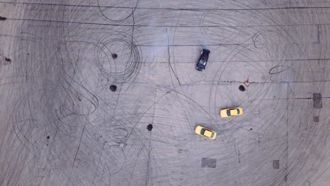 An-Overhead-Aerial-View-of-a-Black-Drift-Car-Burning-Rubber-on-in-an-Asphalt-Parking-Lot-Joined-by-Two-Yellow-Race-Cars-in-High-Horsepower-Tandem-Drifting