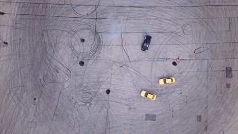 una vista aérea de un coche de deriva negro quemando goma en un estacionamiento de asfalto unido por dos coches de carreras amarillos en tandem de alta potencia a la deriva
