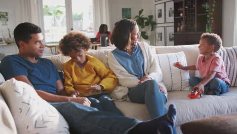 parents and their two children sitting together on sofa, grandparents at a table in the background