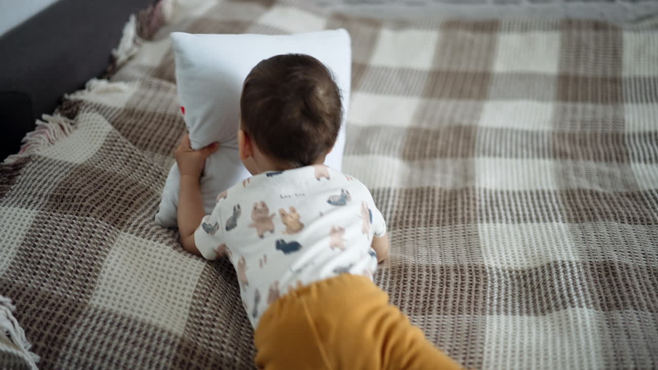 Little happy toddler takes a small pillow and gets off the bed. Adorable baby boy playing at home.