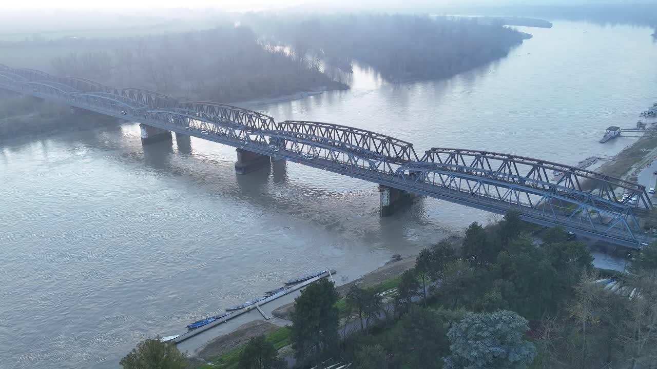 Ripresa aerea distanziata del Ponte di Ferro tra Cremona e Castelvetro Piacentino che mostra il monumentale ponte che attraversa il fiume Po, la foschia nebbiosa invernale e il paesaggio ripariale che si espande con l'altitudine