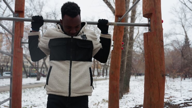 Young man in winter jacket chewing gum jumps to grab an iron bar for pull-ups in a snowy park, dropping down after effort, surrounded by bare trees in a serene setting