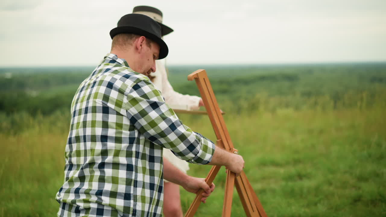 A woman in a white dress, holding a painting brush, watches her husband who is wearing a black hat and plaid shirt as he focuses on setting up a wooden tripod in a scenic grassy field