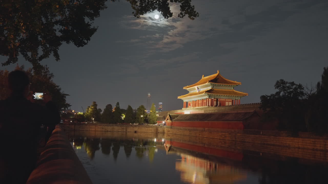 Padiglione illuminato dalla luna vicino al fossato con un fotografo in silhouette che cattura il riflesso di una torre ornata sotto un cielo nuvoloso, caldo bagliore della lanterna, alberi che incorniciano il viale, specchio d'acqua calmo, atmosfera notturna contemplativa.