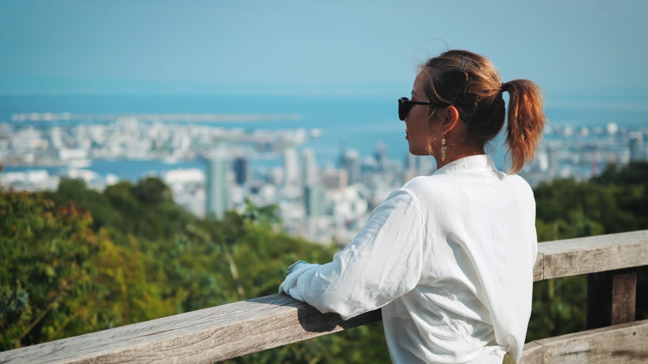 mujer con gafas de sol y camisa blanca mira hacia una ciudad costera desde una barandilla de madera