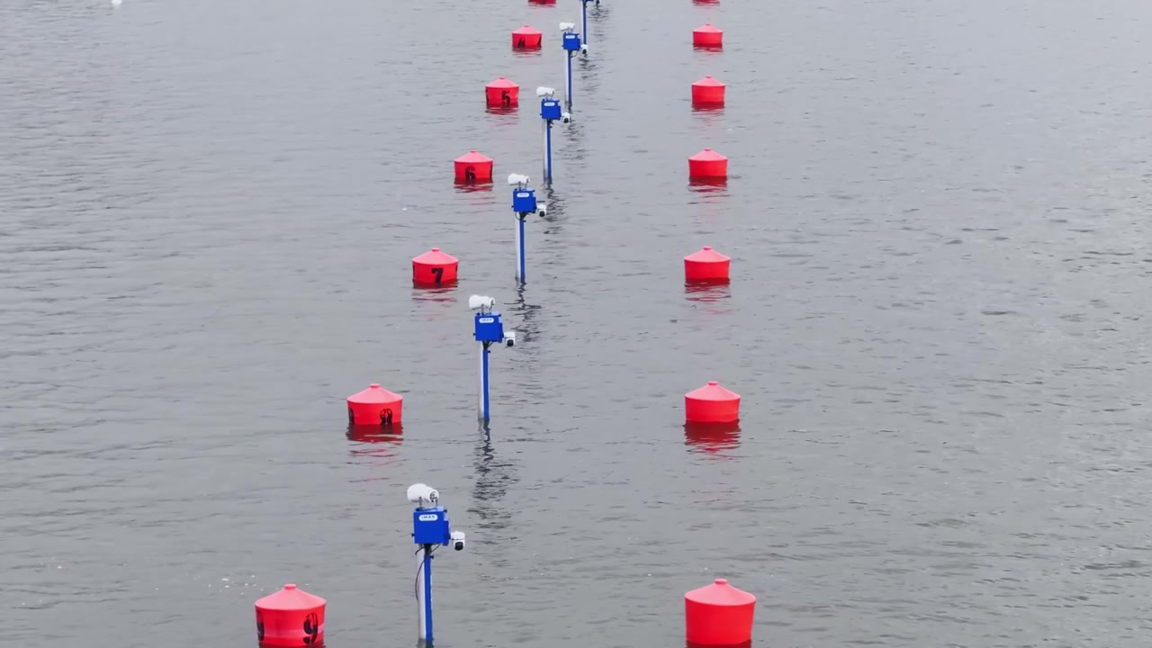 Close-up view of a canoe sprint course lane system, with red floating buoys, blue timing posts, camera boxes and calm grey race water.