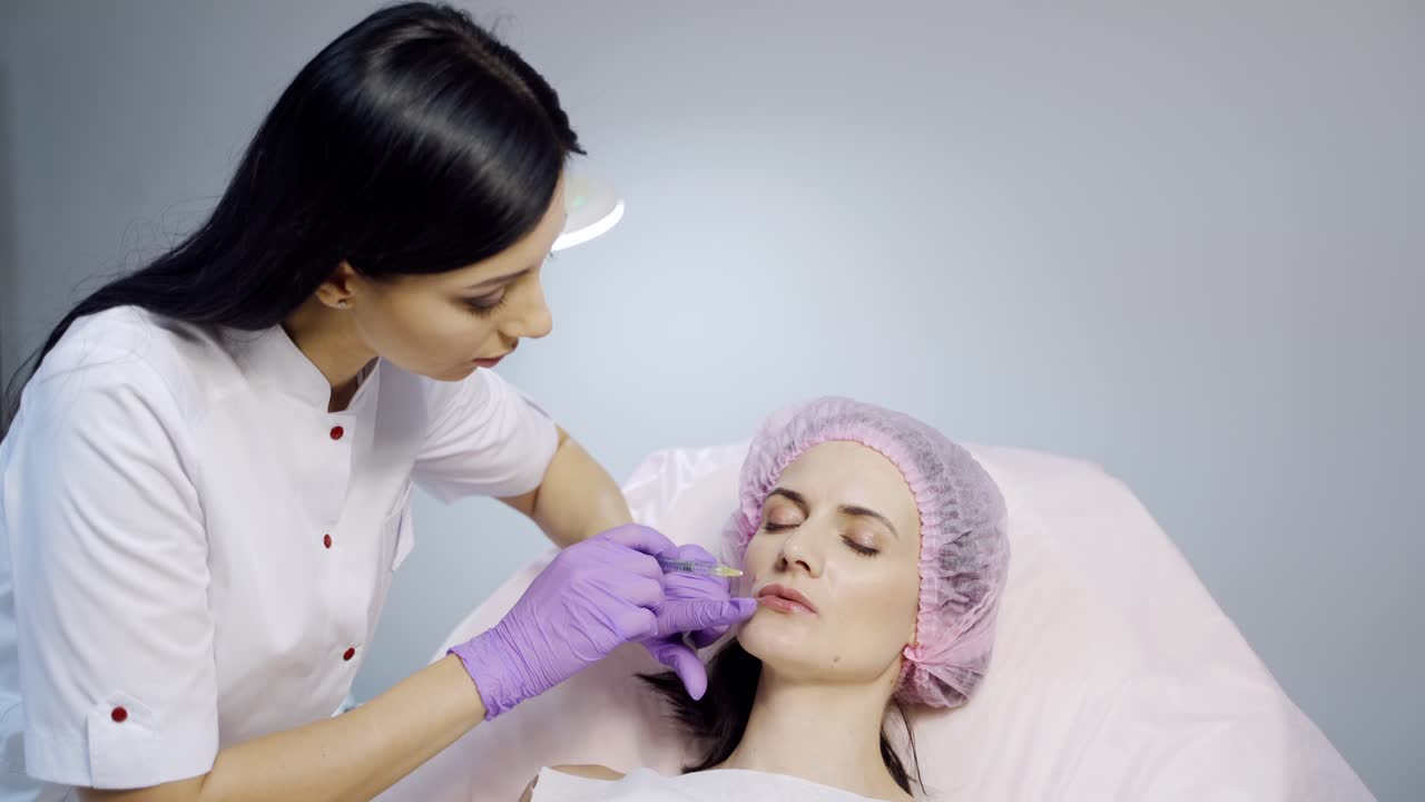 A cosmetologist in a white lab coat performs procedure of lip augmentation for a client in a beauty salon on a light background. Modern methods of cosmetology.