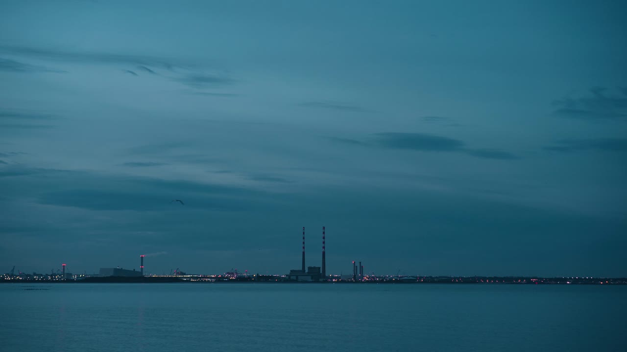 A locked-off shot of Poolbeg Towers and Dublin Port at dusk. The industrial skyline glows with city lights against a deep blue sky, reflecting over calm coastal waters in this atmospheric urban scene.