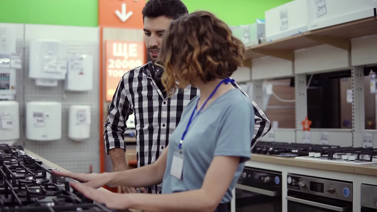 Dark haired, smiling man in plaid shirt with headphones choosing a new modern oven in store. He is planning to buy furniture and