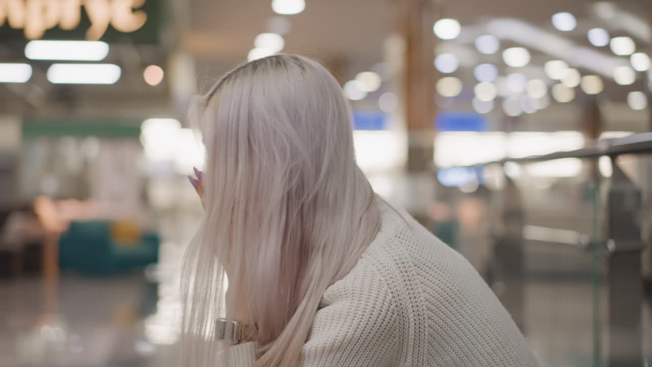 serene woman seated on bench at mall walkway removes phone from ear while listening thoughtfully, wearing white sweater, jeans and boots, glass railing, escalator and decorative holiday lights behind