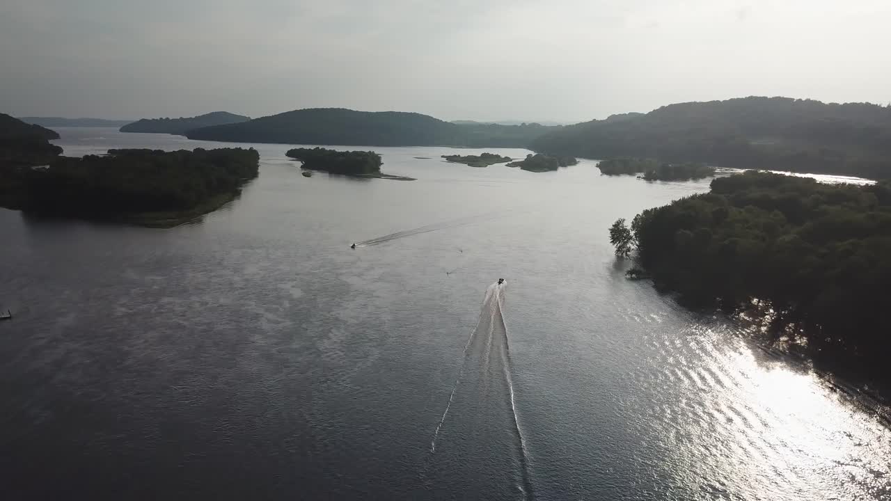 A small boat moves steadily through a wide river channel surrounded by tree-covered islands and distant hills, reflecting soft light on the rippling water.
