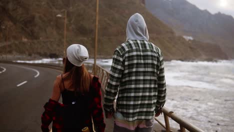 Two-Young-People-Travel-Buddies-Walking-Along-A-Highway,-Against-A-Background-Of-Sea-Coast