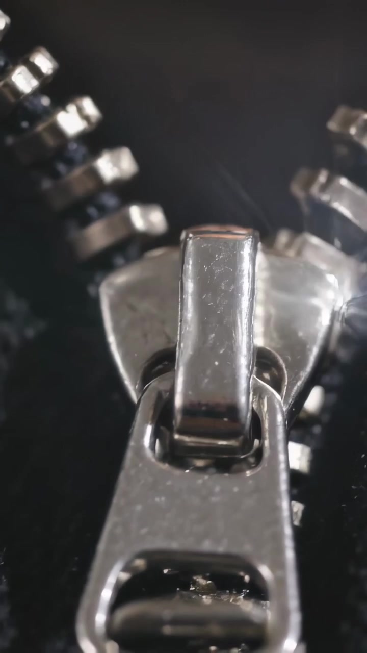 Vidéo verticale: Montrant la tirette et les dents d'une fermeture éclair métallique captant un reflet lumineux sur un tissu sombre