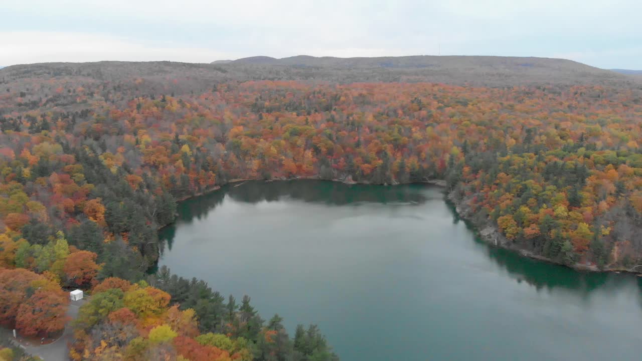 toma aérea inversa desde algunas montañas al otro lado de un gran lago de color azul rodeado por un bosque otoñal de colores brillantes