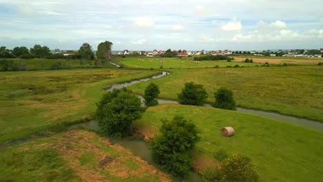 An-aerial-flight-over-green-fields-with-creeks-and-hay-bales,-moving-over-a-row-of-french-style-vacation-homes-onto-the-beach-and-Atlantic-ocean