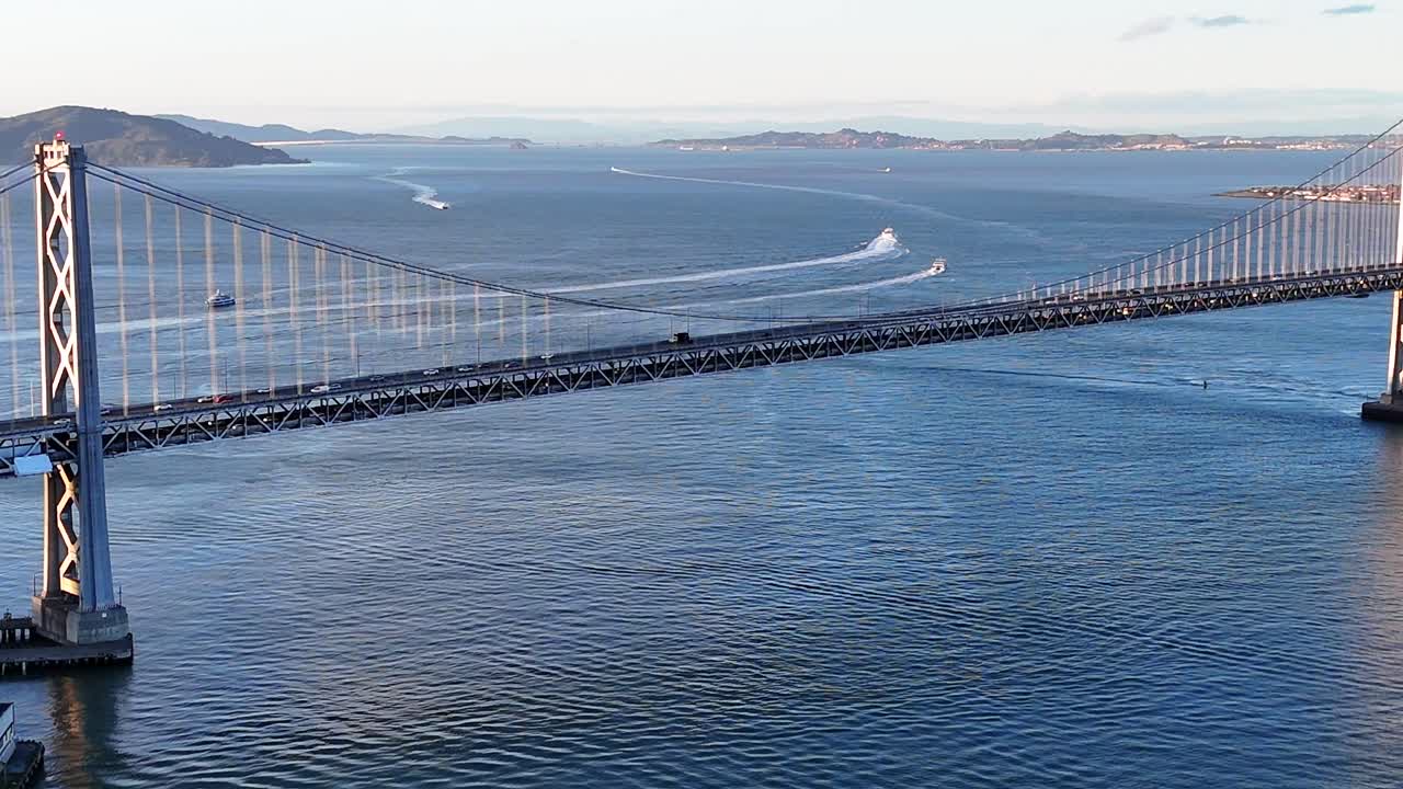 Drone captures a detailed close-up of the San Francisco–Oakland Bay Bridge, showing traffic moving on both upper and lower levels, with boats visible in the San Francisco Bay in the background