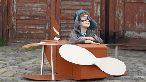 portrait of a cute little boy with red hair in hat and glasses sitting outdoor at wooden model of plane and smiling at the camera 1