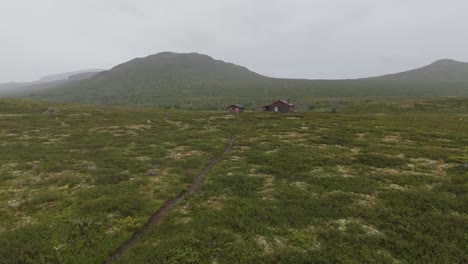 An aerial perspective showcasing a vast grassland under an overcast sky. Small houses are visible in the distance, nestled against a mountainous backdrop.