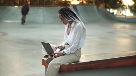 Side-view-of-beautiful,-elegant-girl-with-black-and-white-dreadlocks-sitting-on-parapet-in-local-skatepark-working-on-laptop