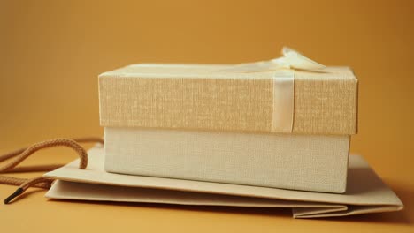 a gift box wrapped in beige paper with a white ribbon and a brown shopping bag on a tan background