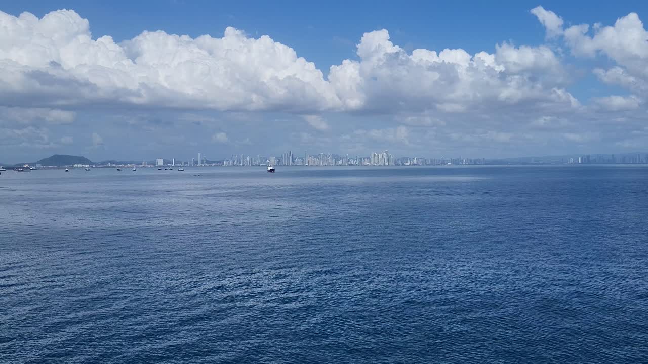 Distant aerial view of Panama City skyline across calm ocean water under a blue sky with clouds.