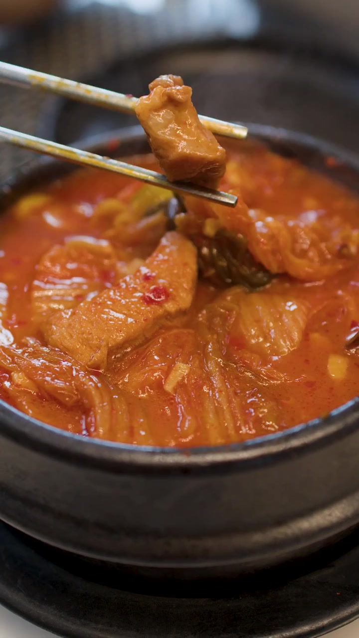 Gambar Jarak Dekat Kimchi Jjigae yang Mengepul dengan Daging Babi dan Tahu yang Sedang Dimakan dengan Sumpit Logam