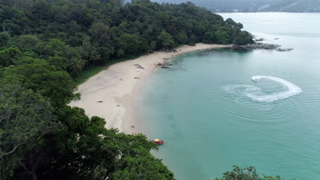 Die-Wunderbare-Aussicht-Auf-Die-üppig-Grüne-Insel-In-Phuketthailand,-Umgeben-Von-Tiefblauem-Ozean-An-Einem-Sonnigen-Tag---Jet-Ski,-Der-Sich-In-Der-Nähe-Der-Küste-Bewegt---Luftaufnahme