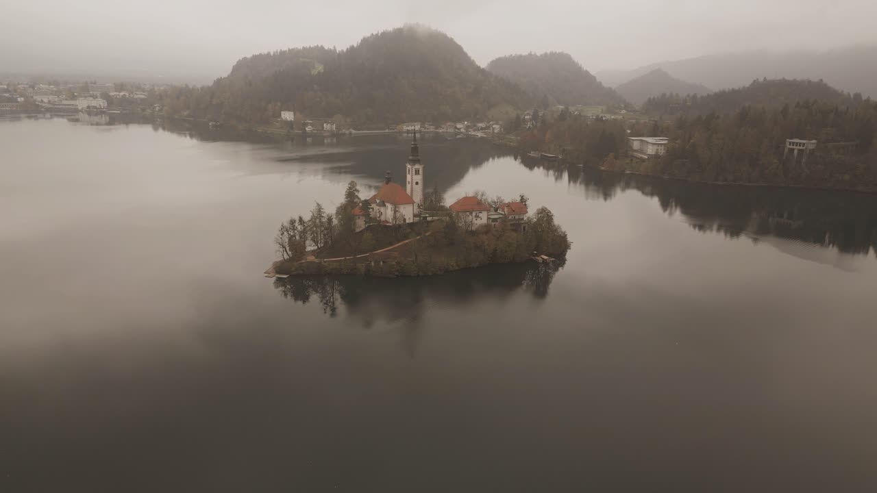 Drone view over Lake Bled, Slovenia, on a cloudy autumn day: misty fog drifts over the tranquil lake, surrounded by colorful autumn trees, with Bled Castle perched on a hill and distant mountains
