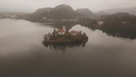 drone view over lake bled, slovenia, on a cloudy autumn day: misty fog drifts over the tranquil lake, surrounded by colorful autumn trees, with bled castle perched on a hill and distant mountains