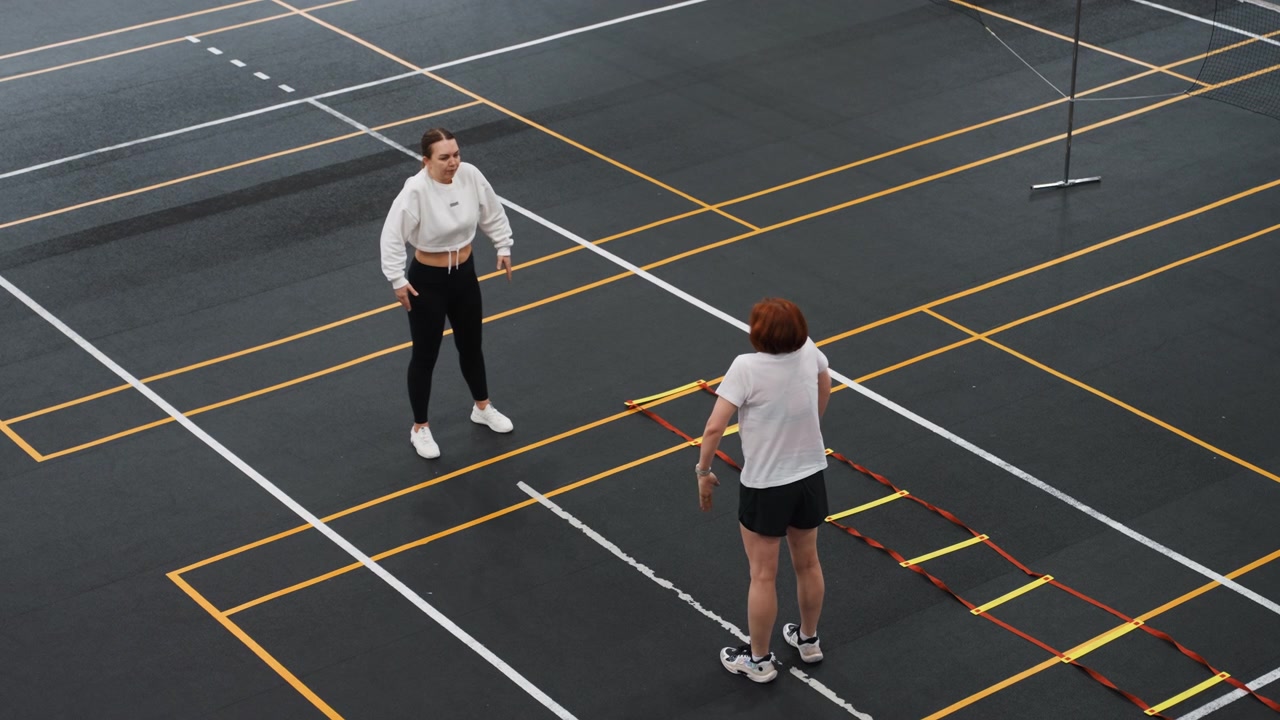 Court badminton footwork practice session, coach demonstrating ladder steps while junior athlete observes and repeats, concentrated technical training, clear court markings and net support visible