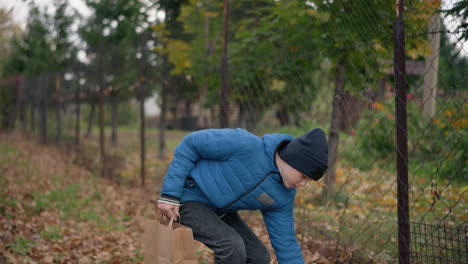 russian boy in blue jacket and beanie bends down to pick yellow leaf from ground and places it into paper bag amidst vibrant autumn foliage and fallen leaves next to rustic outdoor fence