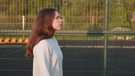 woman walks by fence of city stadium. young lady goes to fitness class to improve physical health thinking about problems at sunset. feeling of solitude