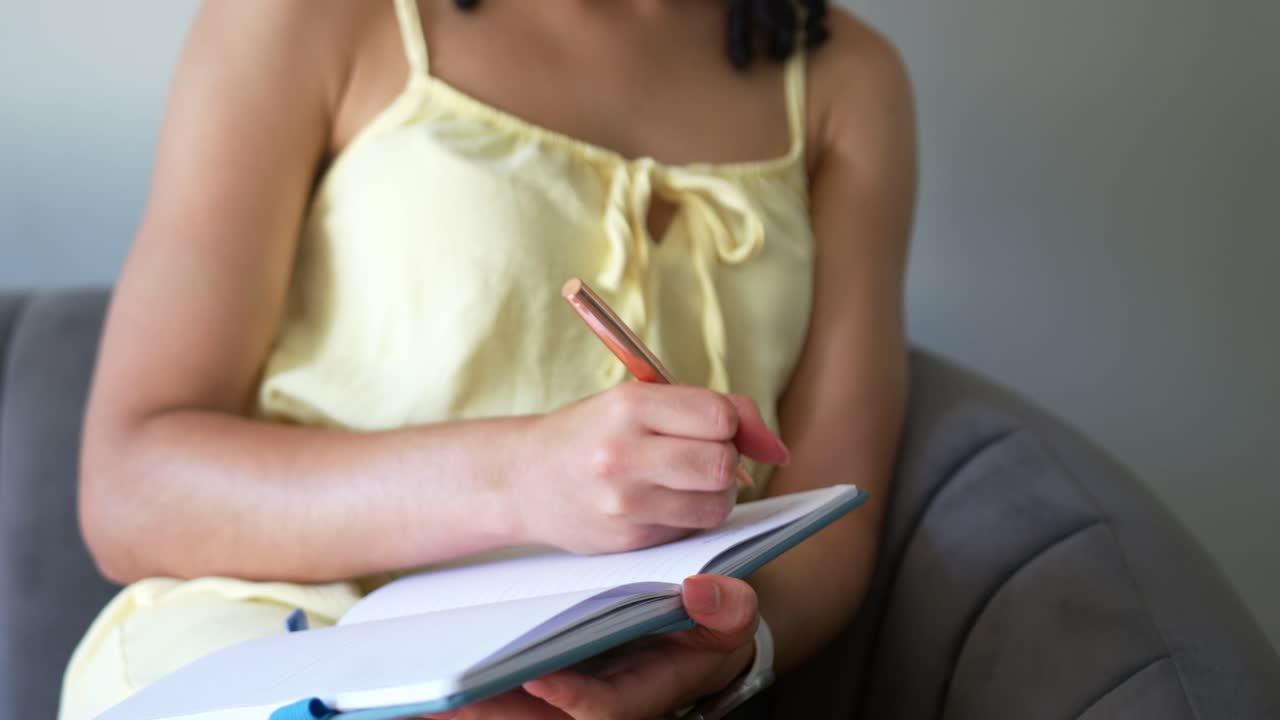 Female holding notebook on lap and lifting rose-gold pen, writing notes in home gray armchair