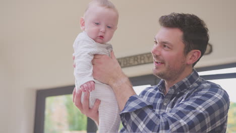 Working-father-using-laptop-at-home-whilst-playing-with-smiling-baby-son-lifting-him-in-the-air---shot-in-slow-motion