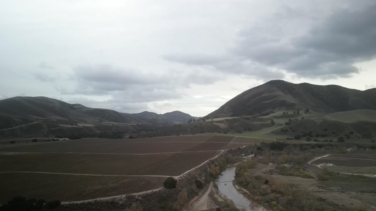 Aerial view of arroyo seco river flowing through agricultural fields, with mountains and cloudy sky in the background, near greenfield, california, drone orbiting shot