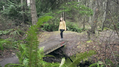 a young woman wearing a bright yellow jacket walks over a small bridge in a green mossy forest, wide panning shot from behind a fern