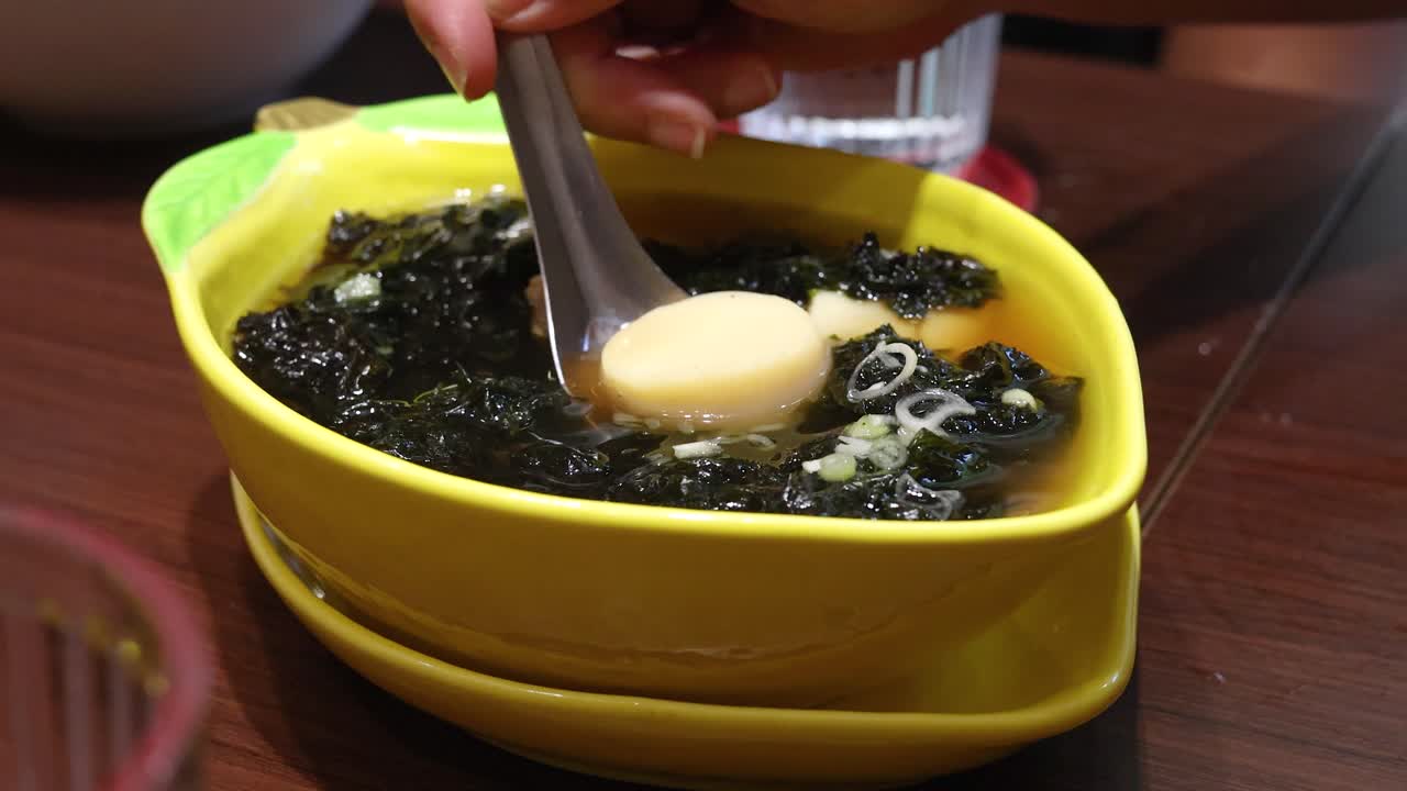 Hand uses spoon to lift pork ball and seaweed from yellow bowl of Thai soup on wooden table, under warm indoor lighting with shallow depth of field
