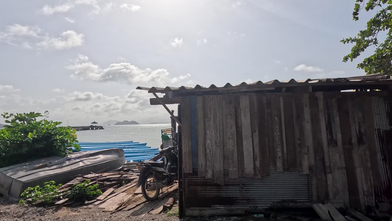 Camera moves along a shaded tropical shoreline, revealing rustic wooden huts, scattered belongings, and tranquil sea views under bright natural daylight