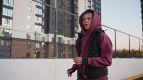 exercise lover wearing hoodie jogs around curved outdoor court lined with barbed wire fence under pastel dusk sky, silhouetted against urban backdrop of apartments and industrial building at sunset