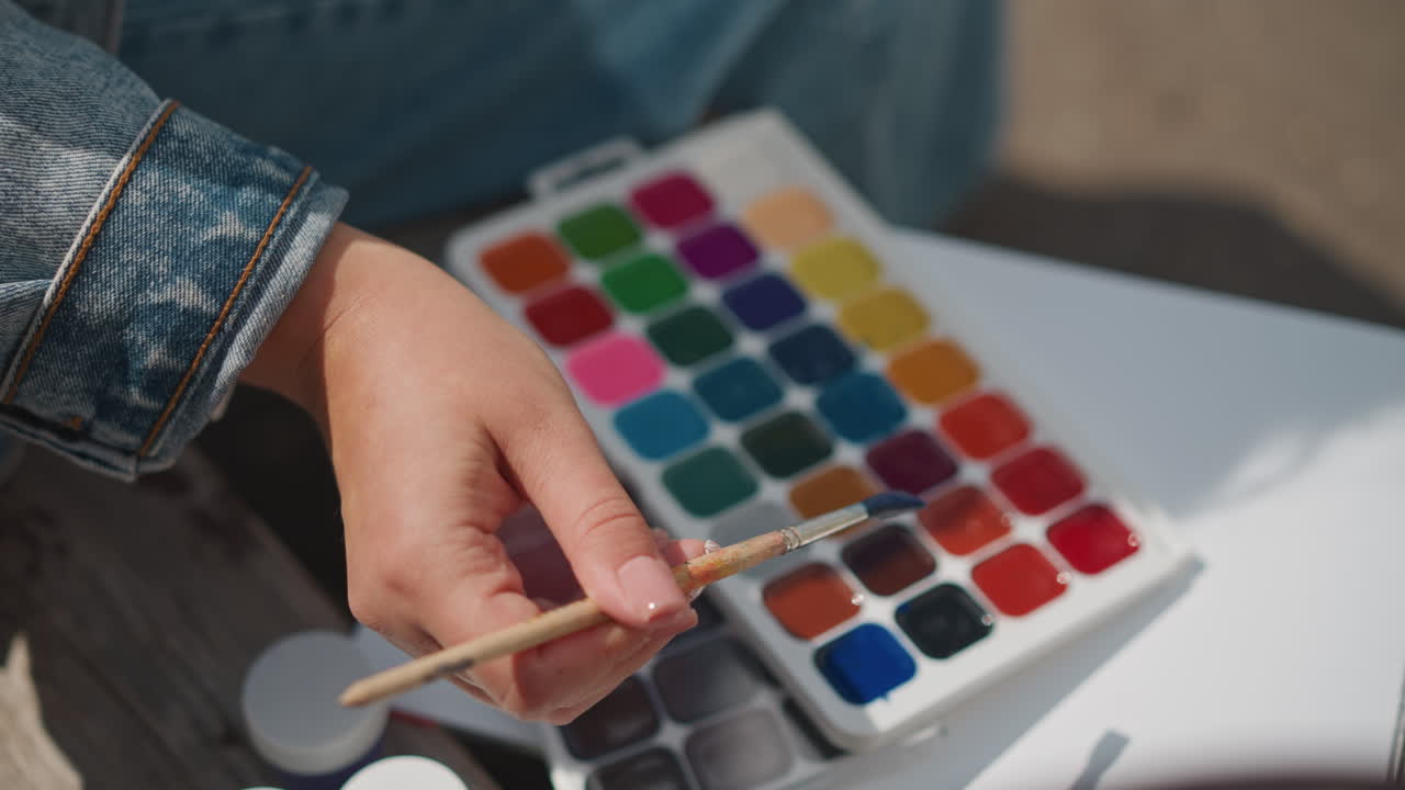 Primer plano de manos mezclando paleta de acuarela azul, manga de chaqueta vaquera, bandeja de mezcla blanca sobre cuaderno de bocetos, la luz del sol resalta las pastillas vibrantes, pincel sumergiéndose en pigmento, preparación creativa tranquila al aire libre