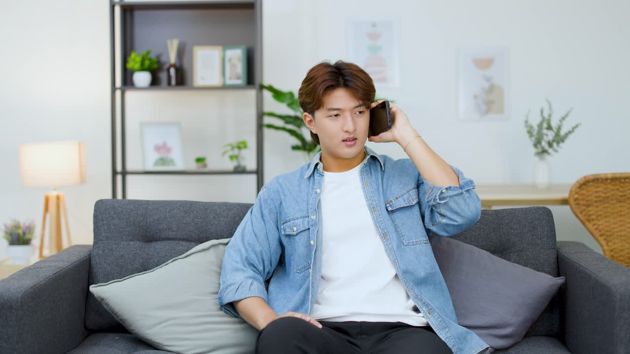 Frustrated East Asian man arguing on smartphone while sitting on a living room sofa