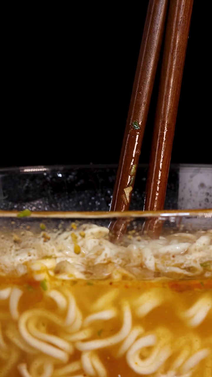 Bacchette marroni mescolano noodles istantanei fumanti in una ciotola di vetro trasparente piena di brodo, sotto una brillante illuminazione da studio su uno sfondo nero, vista ravvicinata.