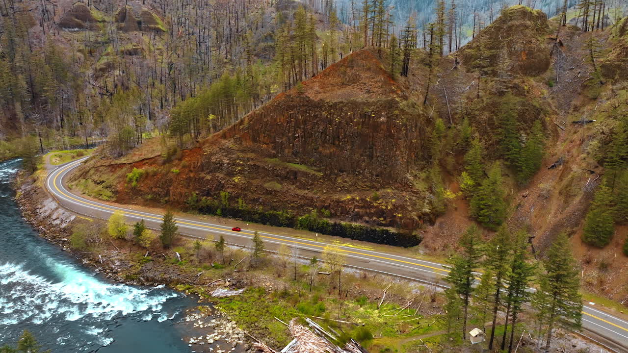 Flying closer to the highway at the foot of the mountains. Red car rides by the road along the rock. Oregon nature.