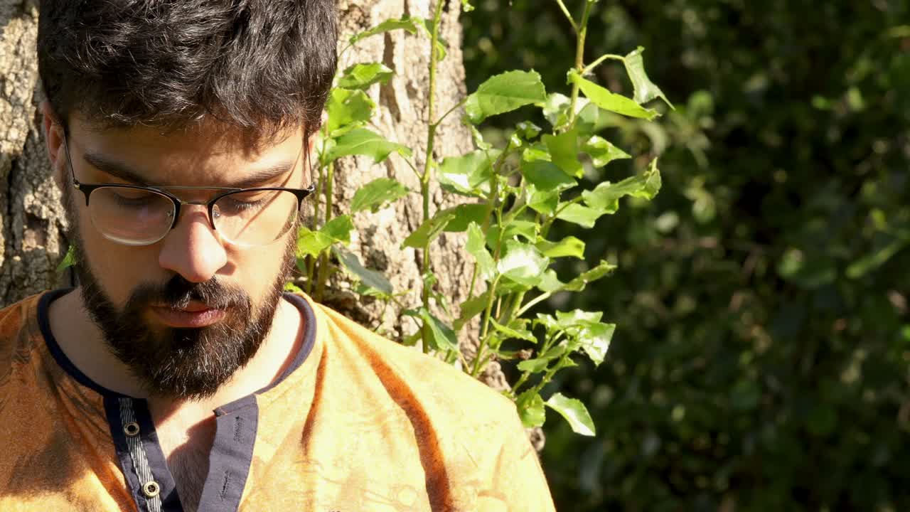 Young handsome man with beard, glasses and black hair with his back on a tree. Outdoors video in 4k.