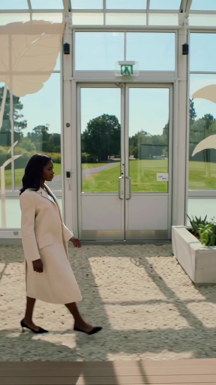 Vertical video: Walking woman in beige coat entering left, crossing greenhouse toward exit sign