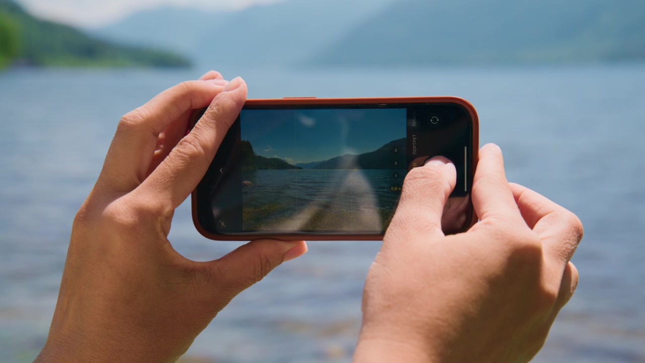 Mãos seguram smartphone capturando uma vista panorâmica do lago. A pessoa se concentra intensamente na tela. A beleza serena da natureza preenche o enquadramento. Ideal para conteúdo de viagem, tecnologia ou com tema de natureza.