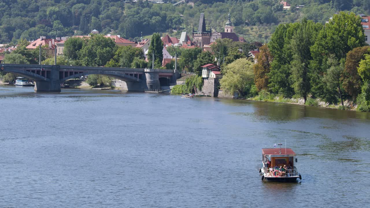 A small passenger boat moves upriver toward a bridge, sunny day, Prague cityscape background