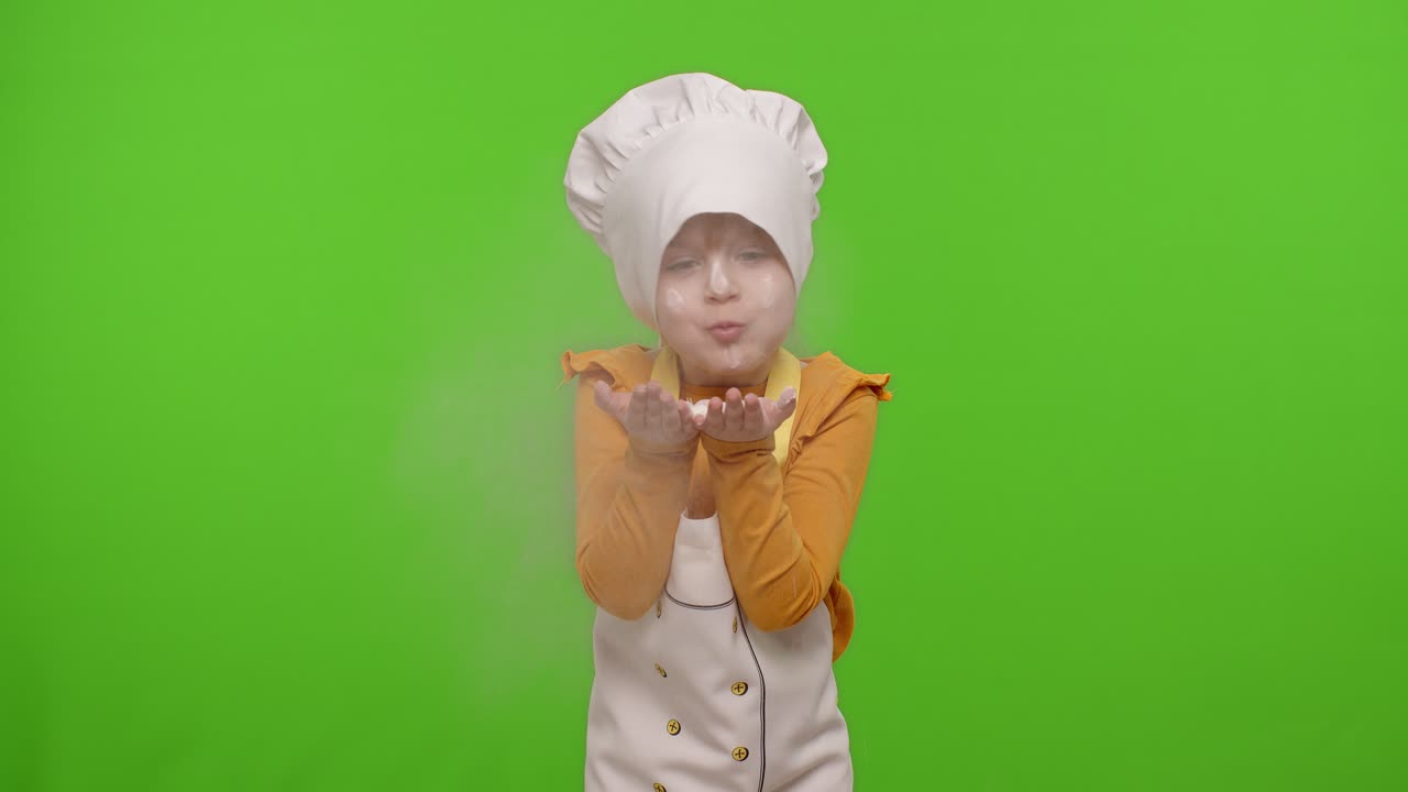 Bambina vestita da cuoca professionista che soffia farina dalle mani verso la telecamera su chroma key.