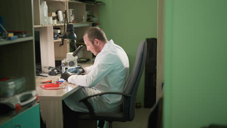 una vista cercana de un técnico en una bata de laboratorio blanca relajándose en su asiento mientras examina cuidadosamente un circuito bajo un microscopio en su laboratorio, con la pared pintada de verde y un estante al lado